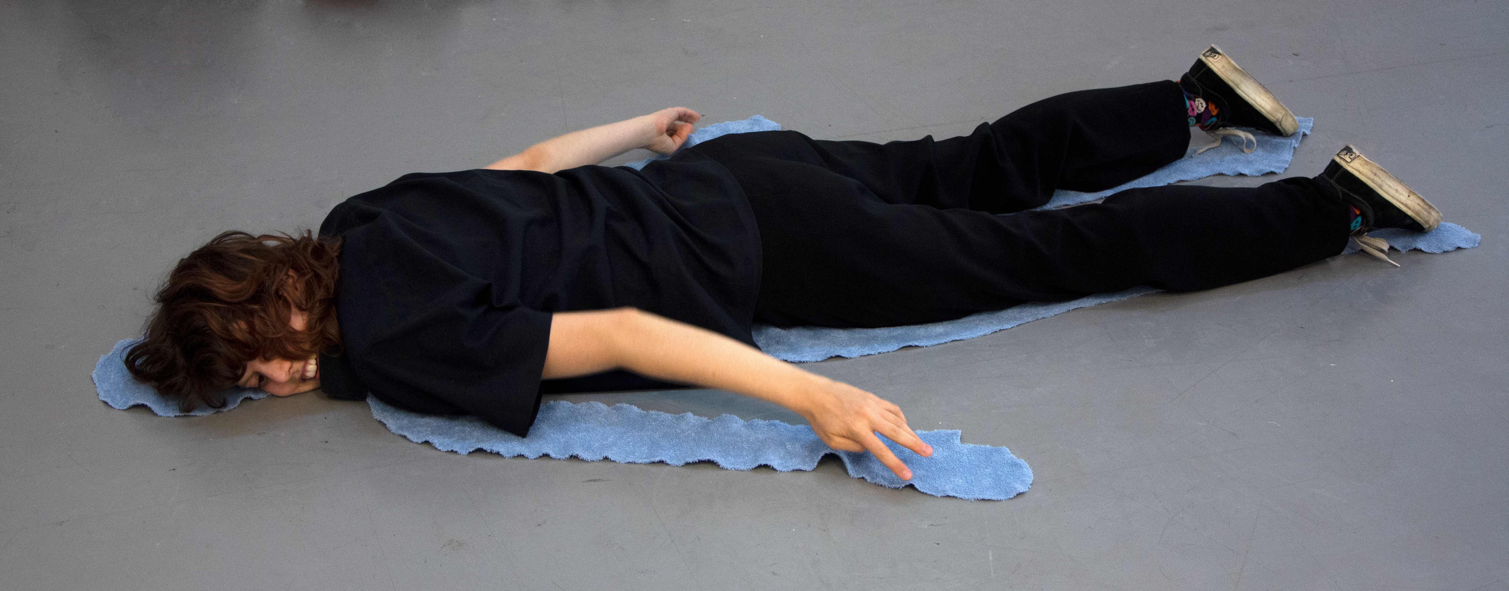 Towel in the shape of a human body on the floor. On top lays the artist, hugging the towel in a humorous  manner.